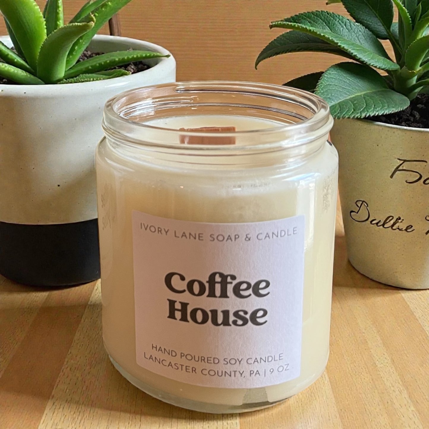 Candle labeled 'Coffee House' by Ivory Lane Soap & Candle on a wooden surface with plants in the background.