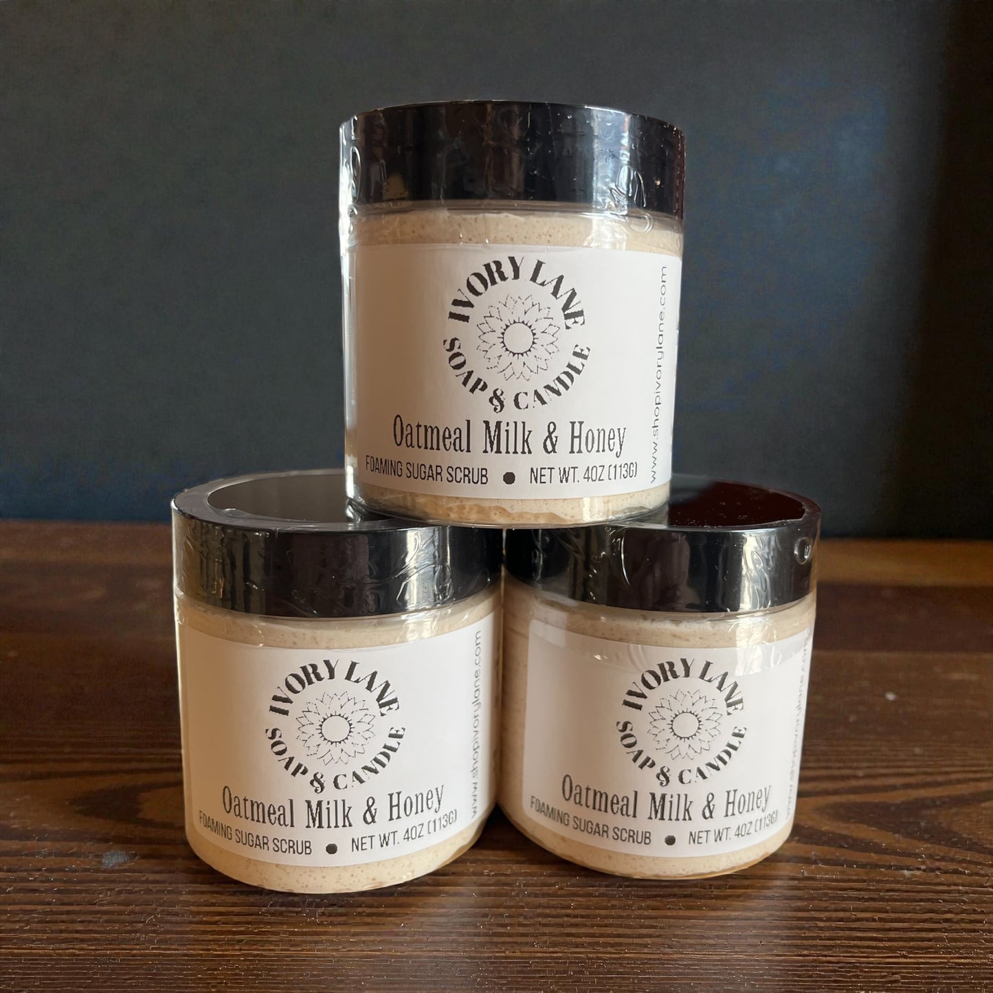 Three jars of Oatmeal Milk & Honey scrub from Ivory Lane Soap & Candle stacked on a wooden surface with a dark background.